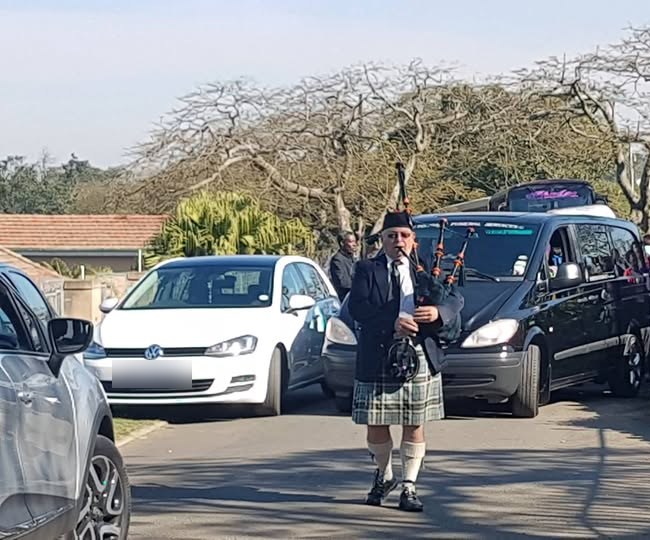 A piper in a kilt leading a funeral procession along a Tongaat street, followed by the funeral vehicle